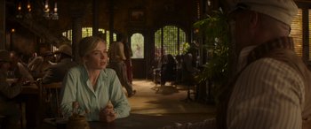Movie still from “Jungle Cruise” (2021), directed by Jaume Collet-Serra – A woman sitting at a table in front of a group of people; Medium shot, Over the shoulder angle
