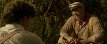 Movie still from “Jungle Cruise” (2021), directed by Jaume Collet-Serra – A man wearing a white hat sitting in front of a tree; Medium shot, Over the shoulder angle