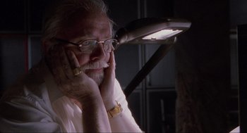 Movie still from “Jurassic Park” (1993), directed by Steven Spielberg – An older man with glasses is sitting in a dimly - lit room; Close Up shot, Low angle