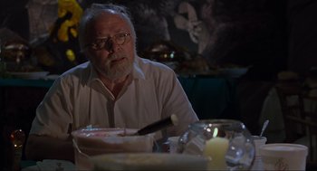 Movie still from “Jurassic Park” (1993), directed by Steven Spielberg – An older man sitting at a table in front of a cake; Medium shot, Over the shoulder angle