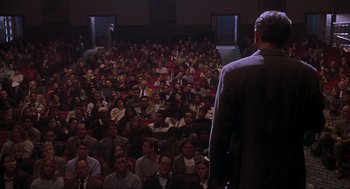 Movie still from “Jurassic Park III” (2001), directed by Joe Johnston – A man standing in front of a crowd of people in a theater; Wide shot, High angle