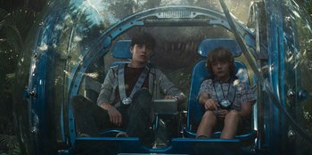 Movie still from “Jurassic World” (2015), directed by Colin Trevorrow – Two young boys sitting in a vehicle with a dinosaur in the background; Medium shot, Over the shoulder angle