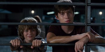 Movie still from “Jurassic World” (2015), directed by Colin Trevorrow – Two young people sitting next to each other in front of a fence; Medium shot, Low angle