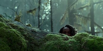 Movie still from “Jurassic World Dominion” (2022), directed by Colin Trevorrow – A person with dreadlocks hiding behind a tree trunk; Medium shot, Low angle