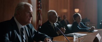 Movie still from “Jurassic World: Fallen Kingdom” (2018), directed by J.A. Bayona – Two men in business suits sitting in front of microphones in front of american flags; Medium shot, Low angle