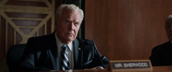 Movie still from “Jurassic World: Fallen Kingdom” (2018), directed by J.A. Bayona – An older man wearing a suit and tie sitting in front of a microphone; Close Up shot, Low angle