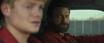 Movie still from “Kandahar” (2023), directed by Ric Roman Waugh – A man in a red shirt and a woman in a red shirt in the back of a car; Close Up shot, Over the shoulder angle