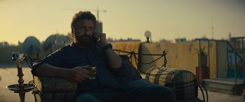 Movie still from “Kandahar” (2023), directed by Ric Roman Waugh – A man sitting on a couch talking on a phone; Medium shot, Over the shoulder angle