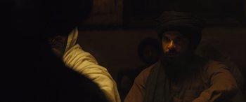 Movie still from “Kandahar” (2023), directed by Ric Roman Waugh – A man with a beard and a turban in a dark room; Medium shot, Over the shoulder angle