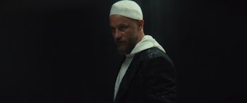 Movie still from “Kandahar” (2023), directed by Ric Roman Waugh – A man with a beard wearing a white hat; Close Up shot, Low angle