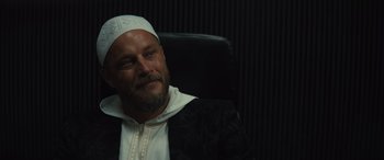 Movie still from “Kandahar” (2023), directed by Ric Roman Waugh – A man with a beard wearing a white hat; Close Up shot, Low angle