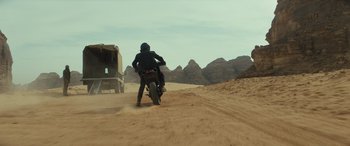 Movie still from “Kandahar” (2023), directed by Ric Roman Waugh – A man riding a motorcycle on a dirt road near mountains; Wide shot, Low angle