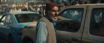 Movie still from “Kandahar” (2023), directed by Ric Roman Waugh – An older man in a red hat is standing in the middle of a busy street; Medium shot, Low angle