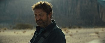 Movie still from “Kandahar” (2023), directed by Ric Roman Waugh – A man with a beard is standing in the desert; Close Up shot, Over the shoulder angle