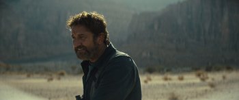 Movie still from “Kandahar” (2023), directed by Ric Roman Waugh – A man with a beard is standing in the desert; Close Up shot, Over the shoulder angle