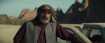 Movie still from “Kandahar” (2023), directed by Ric Roman Waugh – An older man in a turban standing next to a car; Medium shot, Over the shoulder angle