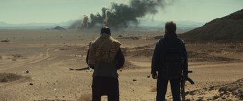 Movie still from “Kandahar” (2023), directed by Ric Roman Waugh – Two men standing in the middle of the desert looking at smoke; Wide shot, Over the shoulder angle