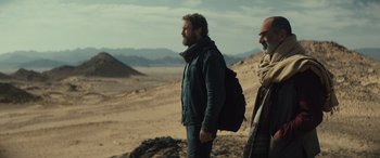 Movie still from “Kandahar” (2023), directed by Ric Roman Waugh – A man with a backpack walking on a dirt road; Medium shot, Over the shoulder angle