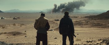 Movie still from “Kandahar” (2023), directed by Ric Roman Waugh – Two men standing in the middle of the desert looking at smoke; Wide shot, Over the shoulder angle