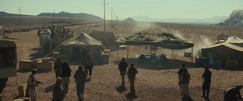 Movie still from “Kandahar” (2023), directed by Ric Roman Waugh – A group of people walking in the dirt near tents and power lines; Extreme Wide shot, High angle