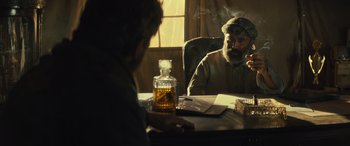 Movie still from “Kandahar” (2023), directed by Ric Roman Waugh – A man sitting at a table smoking a cigarette; Medium shot, Over the shoulder angle