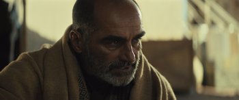 Movie still from “Kandahar” (2023), directed by Ric Roman Waugh – An older man with a beard wrapped in a blanket; Close Up shot, Over the shoulder angle