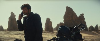 Movie still from “Kandahar” (2023), directed by Ric Roman Waugh – A man standing next to a motorcycle in the desert; Medium shot, Over the shoulder angle
