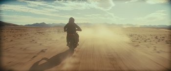 Movie still from “Kandahar” (2023), directed by Ric Roman Waugh – A person riding a motorcycle on a dusty road; Wide shot, Low angle