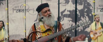 Movie still from “Khufiya” (2023), directed by Vishal Bhardwaj – An old man with a beard and a beard band is holding a guitar; Medium shot, Low angle