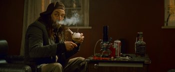 Movie still from “Kick-Ass” (2010), directed by Matthew Vaughn – A man sitting on a table smoking a pipe and holding an apple in his hand; Medium shot, High angle