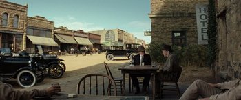 Movie still from “Killers of the Flower Moon” (2023), directed by Martin Scorsese – Two men sitting at an outdoor table in a western town; Wide shot, Over the shoulder angle