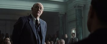 Movie still from “Killers of the Flower Moon” (2023), directed by Martin Scorsese – An older man in a suit and tie in front of a group of people; Medium shot, Over the shoulder angle