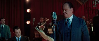 Movie still from “Killers of the Flower Moon” (2023), directed by Martin Scorsese – A man in a suit and tie is holding a microphone; Medium shot, Over the shoulder angle