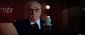 Movie still from “Killers of the Flower Moon” (2023), directed by Martin Scorsese – An older man in a suit and tie holding a microphone; Close Up shot, Over the shoulder angle