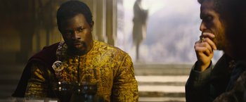 Movie still from “King Arthur: Legend of the Sword” (2017), directed by Guy Ritchie – A man in a yellow and black jacket is holding a cup; Medium shot, Over the shoulder angle