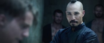 Movie still from “King Arthur: Legend of the Sword” (2017), directed by Guy Ritchie – A man with a shaved head is looking at the camera while another man looks on; Close Up shot, Over the shoulder angle