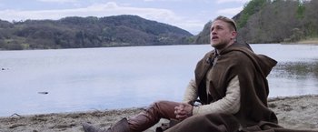Movie still from “King Arthur: Legend of the Sword” (2017), directed by Guy Ritchie – A man sitting next to a body of water; Medium shot, Low angle