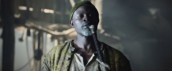 Movie still from “King Arthur: Legend of the Sword” (2017), directed by Guy Ritchie – A black man with a beard and a green hat is smoking a pipe; Close Up shot, Low angle