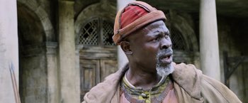 Movie still from “King Arthur: Legend of the Sword” (2017), directed by Guy Ritchie – An older man with a beard and a red hat is standing in front of a building; Close Up shot, Low angle