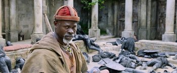 Movie still from “King Arthur: Legend of the Sword” (2017), directed by Guy Ritchie – An older man wearing a red hat standing in front of statues; Medium shot, Over the shoulder angle