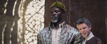 Movie still from “King Arthur: Legend of the Sword” (2017), directed by Guy Ritchie – A man with a green hat is standing in front of a wall; Medium shot, Low angle