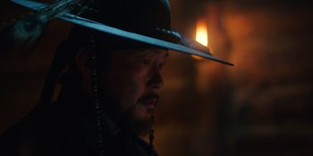 Movie still from “Kingdom: Ashin of the North” (2021), directed by Kim Seong-hun – A person wearing a hat with a candle in the background; Close Up shot, Over the shoulder angle