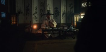 Movie still from “Kingdom: Ashin of the North” (2021), directed by Kim Seong-hun – A man sitting at a table with a lit candle in front of him; Wide shot, Low angle