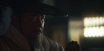 Movie still from “Kingdom: Ashin of the North” (2021), directed by Kim Seong-hun – An old man with a beard wearing a black hat; Close Up shot, Over the shoulder angle