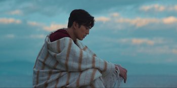 Movie still from “Knights of the Zodiac” (2023), directed by Tomasz Baginski – A man wrapped in a blanket sitting on the beach; Medium shot, Low angle