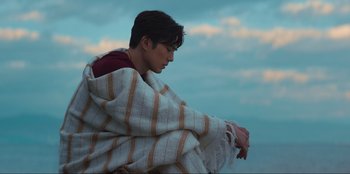 Movie still from “Knights of the Zodiac” (2023), directed by Tomasz Baginski – A man wrapped in a blanket sitting on the ground; Medium shot, Low angle