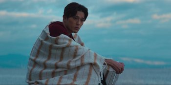 Movie still from “Knights of the Zodiac” (2023), directed by Tomasz Baginski – A man wrapped in a blanket sitting on a chair; Medium shot, Low angle