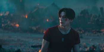 Movie still from “Knights of the Zodiac” (2023), directed by Tomasz Baginski – A young man in a red t - shirt is standing in front of fire; Medium shot, Low angle