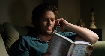 Movie still from “Knocked Up” (2007), directed by Judd Apatow – A man sitting in a chair talking on a cell phone while reading a book; Close Up shot, High angle