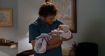 Movie still from “Knocked Up” (2007), directed by Judd Apatow – A man holding a baby in his arms in a living room; Medium shot, Over the shoulder angle